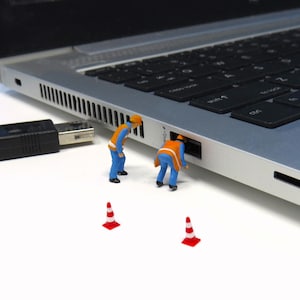 以下が含まれることがあります： 黒いキーボードとUSBケーブルを備えたシルバーのラップトップ。 青とオレンジの制服を着た2人のミニチュア建設作業員が、ラップトップのポートで作業しているようです。 赤と白のトラフィックコーンが2つ、ラップトップの前にあります。