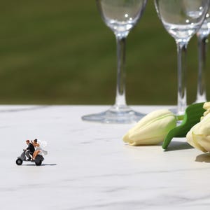 May include: A miniature wedding couple on a scooter, set on a white marble surface. Several wine glasses and white tulips with green leaves are arranged nearby. The background is a blurred green.
