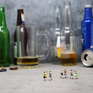 Puede incluir: Una variedad de botellas de cerveza, una jarra y una lata están dispuestas sobre una superficie. Figuras en miniatura con trajes tradicionales alemanes bailan en primer plano. Las botellas son de varios colores, incluyendo verde y azul.