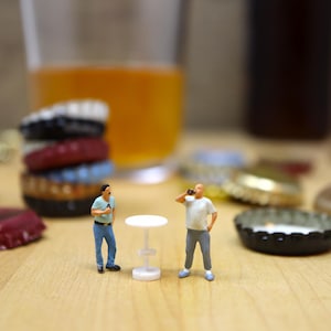 Peut inclure: Une scène miniature avec deux petites figurines près d'une petite table blanche. Une pile de capsules de bouteilles colorées et un verre de liquide ambré sont en arrière-plan. Plusieurs capsules sont éparpillées sur la surface en bois.