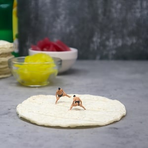May include: A humorous image showing two tiny sumo wrestlers on a large tortilla. In the background are bowls of yellow and red food, and a stack of tortillas. The scene is set on a grey surface.