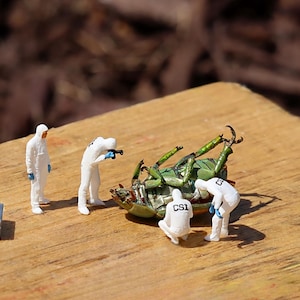 Puede incluir: Escena en miniatura de un insecto verde examinado por cuatro figuras con trajes blancos de CSI sobre una superficie de madera. También hay dos pequeñas cajas grises. El fondo está borroso.
