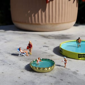 May include: Miniature figures enjoying a sunny day at the beach. A woman relaxes in a beach chair, while others swim in bottle cap pools. A large, beige pot is in the background.