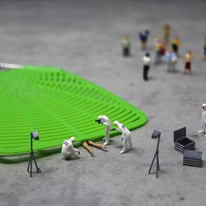 Puede incluir: Una escena del crimen en miniatura con investigadores forenses con trajes blancos examinando pruebas. Una gran matamoscas verde está en el fondo. Pequeñas figuras de personas están a lo lejos. También hay dos cajas negras y dos pequeñas luces.