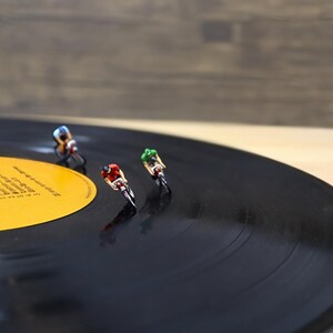 Puede incluir: Ciclistas en miniatura compitiendo en un disco de vinilo negro. Los ciclistas llevan cascos rojos, verdes y azules. El disco tiene una etiqueta amarilla con texto.