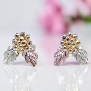 May include: A pair of stud earrings featuring a cluster of gold-colored beads and two silver-colored leaves. The leaves have a pinkish hue. The earrings are set against a soft, blurred background.
