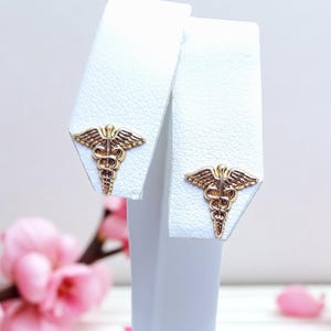 May include: A pair of gold-colored stud earrings, each featuring the Caduceus symbol, displayed on white earring stands. The earrings are set against a soft, blurred background with pink flowers.