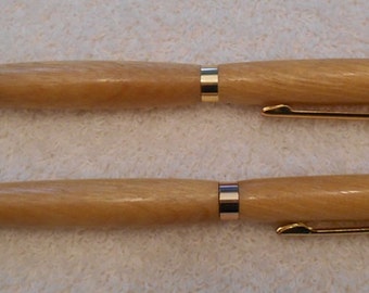 May include: Two wooden pens with gold-colored accents. The pens have a light brown, natural wood grain finish. One pen has a ballpoint tip, and the other has a pencil tip. Both pens have a gold-colored clip.