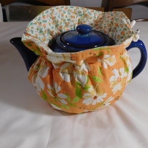 May include: A blue teapot with a floral print tea cozy. The cozy is orange with white flowers and green leaves.