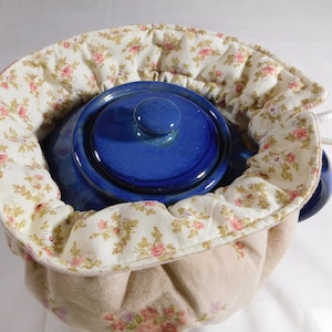 May include: A blue ceramic teapot with a lid inside a beige and floral tea cozy with a drawstring closure.