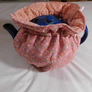 May include: A blue teapot with a floral print tea cozy. The cozy has a drawstring closure and is made of pink fabric with a floral pattern.