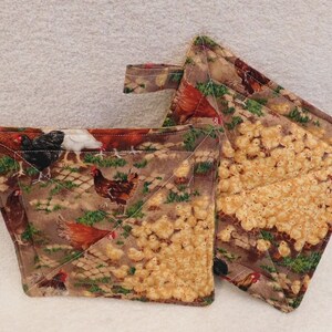 May include: Two brown and white quilted pot holders with a pattern of chickens and chicks. One pot holder has a brown handle.