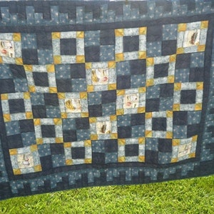 May include: A blue and yellow quilt with a geometric pattern. The quilt is made of squares and rectangles, and it has a border of small squares. The quilt is hanging on a clothesline.