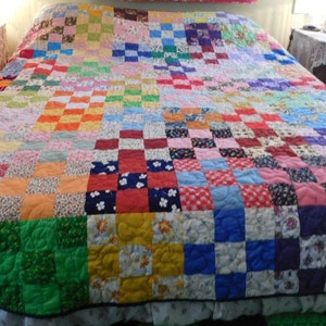 May include: A colourful patchwork quilt, covering a bed. The quilt is made up of numerous squares in a variety of colours and patterns, including floral and solid fabrics. The edges of the quilt are finished with a ruffled trim, adding a decorative touch.
