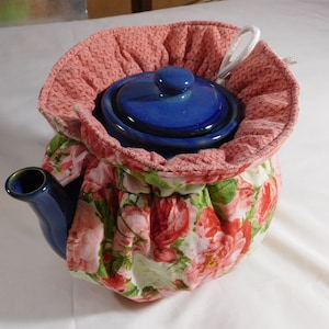 May include: A blue teapot with a floral print tea cozy. The cozy has a pink and white floral pattern with green leaves. The teapot has a lid and a spout.
