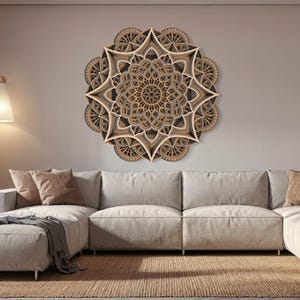 Op de afbeelding: Een groot, ingewikkeld ontworpen houten mandala wanddecor in tinten bruin en beige hangt boven een witte hoekbank in een moderne woonkamer.