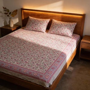 Puede incluir: Una cama doble con estructura y cabecero de madera, cubierta con una colcha estampada con flores y dos fundas de almohada a juego. El estampado presenta flores rosas y grises sobre un fondo crema. Se ve una mesita de noche con un jarrón.