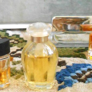 May include: Several small perfume bottles with various designs. One has a black, flower-shaped cap, another a clear, rounded bottle with a clear stopper, and a third with a unique, frosted flower-shaped top. The bottles contain amber-colored liquid.