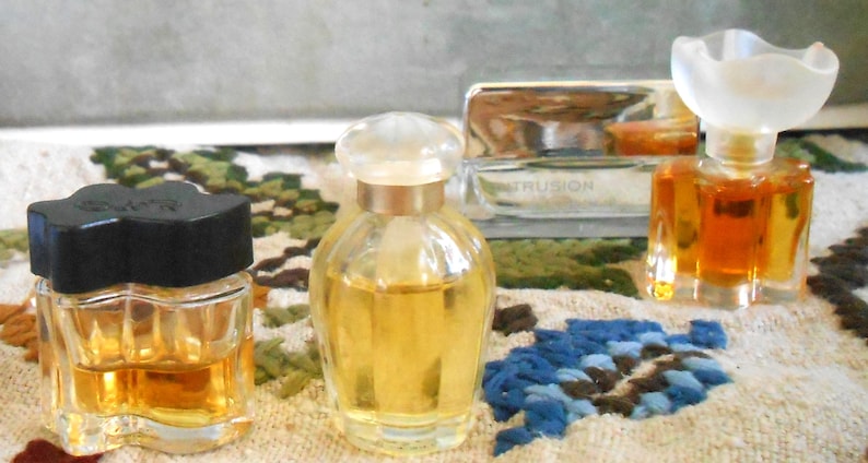 May include: Several small perfume bottles with various designs. One has a black, flower-shaped cap, another a clear, rounded bottle with a clear stopper, and a third with a unique, frosted flower-shaped top. The bottles contain amber-colored liquid.