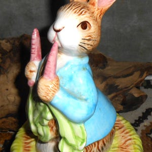 May include: A ceramic figurine of a brown and white rabbit wearing a light blue jacket. The rabbit is standing on a textured yellow base and holding two pinkish-orange carrots with green leafy tops in its front paws.