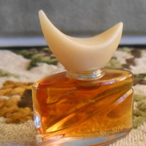 Puede incluir: Un pequeño frasco de perfume vintage con un diseño único. La botella está llena de un líquido de color dorado y tiene un tapón curvo de color crema. La botella tiene forma rectangular con bordes redondeados. El texto "MARC SINA" es visible.