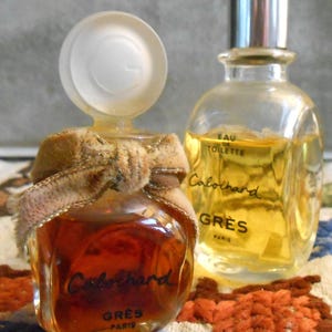 May include: Two vintage perfume bottles. One is a small, round bottle with a brown liquid, a velvet ribbon, and the name "Cabochard" in black script. The other is a taller, clear bottle with a silver cap and the name "Cabochard" and "GRES PARIS".
