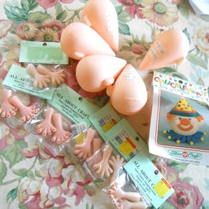 May include: A collection of doll-making supplies, including several peach-colored doll heads, small plastic hands, and a clown head. The hands are packaged in clear plastic, and the clown head is in a Fibre Craft package.