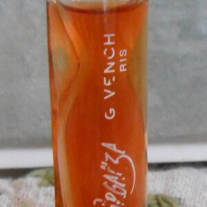 Puede incluir: Un pequeño frasco de perfume con tapón dorado y cuerpo de cristal transparente. El frasco contiene un líquido de color naranja. El texto "Organza" y "GIVENCHY" son visibles en el frasco.