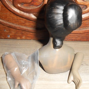 Op de afbeelding: Een porseleinen poppenhoofd met zwart haar in een knot, een naakt torso en twee losse armen. De pop mist haar benen.