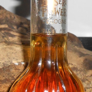 May include: A vintage Desert Flower Cologne bottle with a pale pink cap. The clear glass bottle has a bulbous, ribbed base filled with amber liquid. Gold lettering reads "Desert Flower Cologne".