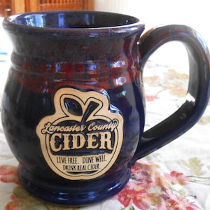 May include: A handcrafted ceramic mug with a dark blue glaze and a red-brown rim. The mug features a large handle and a cream-colored label with the words "Lancaster County CIDER" and the slogan "Live Free. Dine Well. Drink Real Cider."