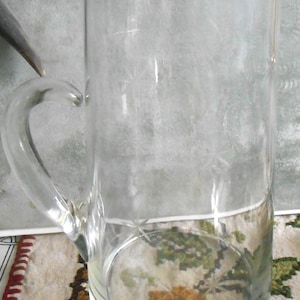 May include: A clear glass beer mug with a silver rim and a curved handle. The glass has etched starburst designs. The mug is approximately 7 inches tall and is sitting on a floral patterned surface.