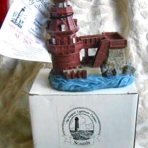May include: A miniature replica of the John Shoal Lighthouse, a red brick lighthouse with a white base and a small wooden bridge. The lighthouse is surrounded by blue water and rocks. The box is labeled "The Original Lighthouse Company, Scaasis, SC-263S".