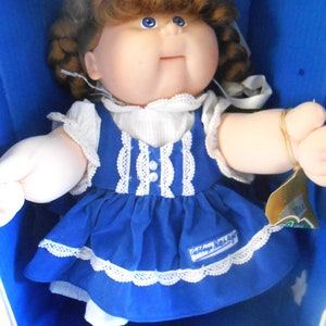 Puede incluir: Una muñeca Cabbage Patch Kids con cabello castaño y ojos azules que lleva un vestido azul y blanco con ribete de encaje blanco. La muñeca está sentada en una caja azul con el logotipo de Cabbage Patch Kids en el vestido.