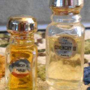 May include: Two small perfume bottles with gold-colored caps. One bottle contains a golden liquid, the other is clear. Both bottles have labels that read "Givenchy III Paris". The bottles are on a patterned surface.