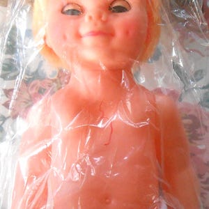 Puede incluir: Una muñeca de plástico con cabello rubio, que lleva un pañal blanco. La muñeca tiene una tez rosada y una sonrisa traviesa. La muñeca está empaquetada en plástico transparente.