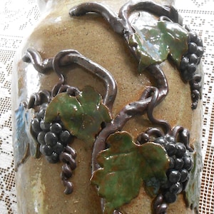 Puede incluir: Un jarrón de cerámica marrón con una superficie texturizada, decorado con un diseño en relieve de vides, hojas y uvas.