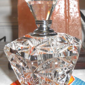 May include: A clear glass bottle with a faceted diamond-shaped stopper. The bottle has a silver metal cap and a faceted design.