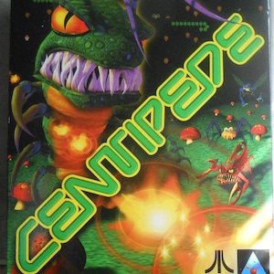 May include: A video game box for Centipede, featuring a green monster with large teeth and orange eyes. The title is in green and yellow. The background shows a green field with mushrooms and other creatures. The Atari and Hasbro Interactive logos are visible.
