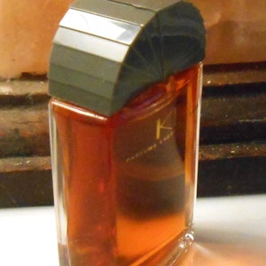 以下が含まれることがあります： ダークグレーの幾何学的なキャップが付いた長方形の香水瓶。ボトルには赤褐色の液体が入っています。「K Parfums Lady」と書かれたラベル。ボトルは透明なガラス製です。