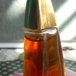 May include: A vintage perfume bottle with a gold-coloured, pyramid-shaped cap. The clear glass bottle contains a reddish-brown liquid. The bottle has a unique, angular design and sits on a colourful floral patterned surface.