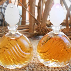 May include: Two clear glass perfume bottles with a swirl design and white leaf-shaped stoppers. The bottles are filled with a golden-colored liquid. One bottle has the text "Creation Ted Lapidus".