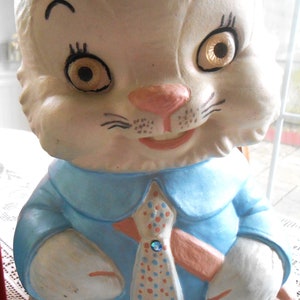 Può includere: Una statuetta di coniglio in ceramica bianca che indossa una camicia blu con una cravatta a pois rossi e bianchi. Il coniglio ha il naso rosa e gli occhi blu.