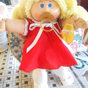 Pode incluir: Uma boneca Cabbage Patch Kid com cabelo de lã loiro, olhos azuis e um vestido vermelho com um laço branco. A boneca está usando meias brancas e sapatos brancos. A boneca está segurando uma chupeta e tem uma etiqueta que diz "The Legend Wilona Esta".