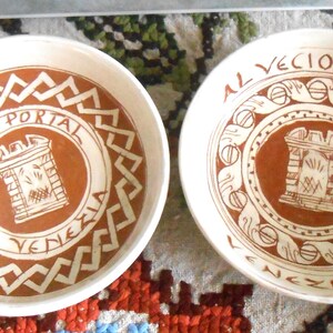 May include: Two white ceramic bowls with a brown and white design. Each bowl features a central brown circle with a drawing of a building and the words "AL VECIO PORTAL VENEZIA" in a circular pattern.