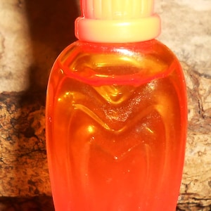 May include: A small, translucent orange plastic bottle with a ribbed, gradient yellow-to-orange cap. The bottle features a molded, heart-shaped indentation on its front surface, creating a smooth, rounded silhouette with a tapered base.