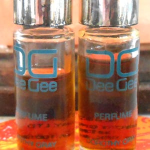 May include: Two small glass bottles of perfume with silver caps. The bottles are filled with a light brown liquid. The labels on the bottles read "Dee Gee Perfume" in blue lettering.