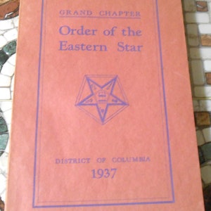 Op de afbeelding: Een roze boekje met de titel "Grand Chapter Order of the Eastern Star" en de tekst "District of Columbia 1937". Op de omslag is een vijfpuntige ster met het nummer 2 in het midden gedrukt.