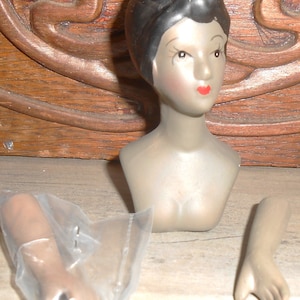Puede incluir: Una cabeza de muñeca de porcelana con cabello negro y un torso gris. A la muñeca le faltan los brazos, que se incluyen por separado en bolsas de plástico.