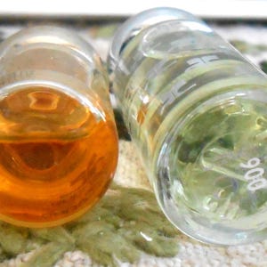 May include: Two clear glass perfume bottles, one filled with amber liquid and the other with a pale green liquid. The bottles are lying on a patterned surface. The bottom of one bottle has the number "906" printed on it.